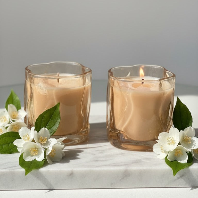 Two lit candles in glass holders with white flowers on a marble surface
