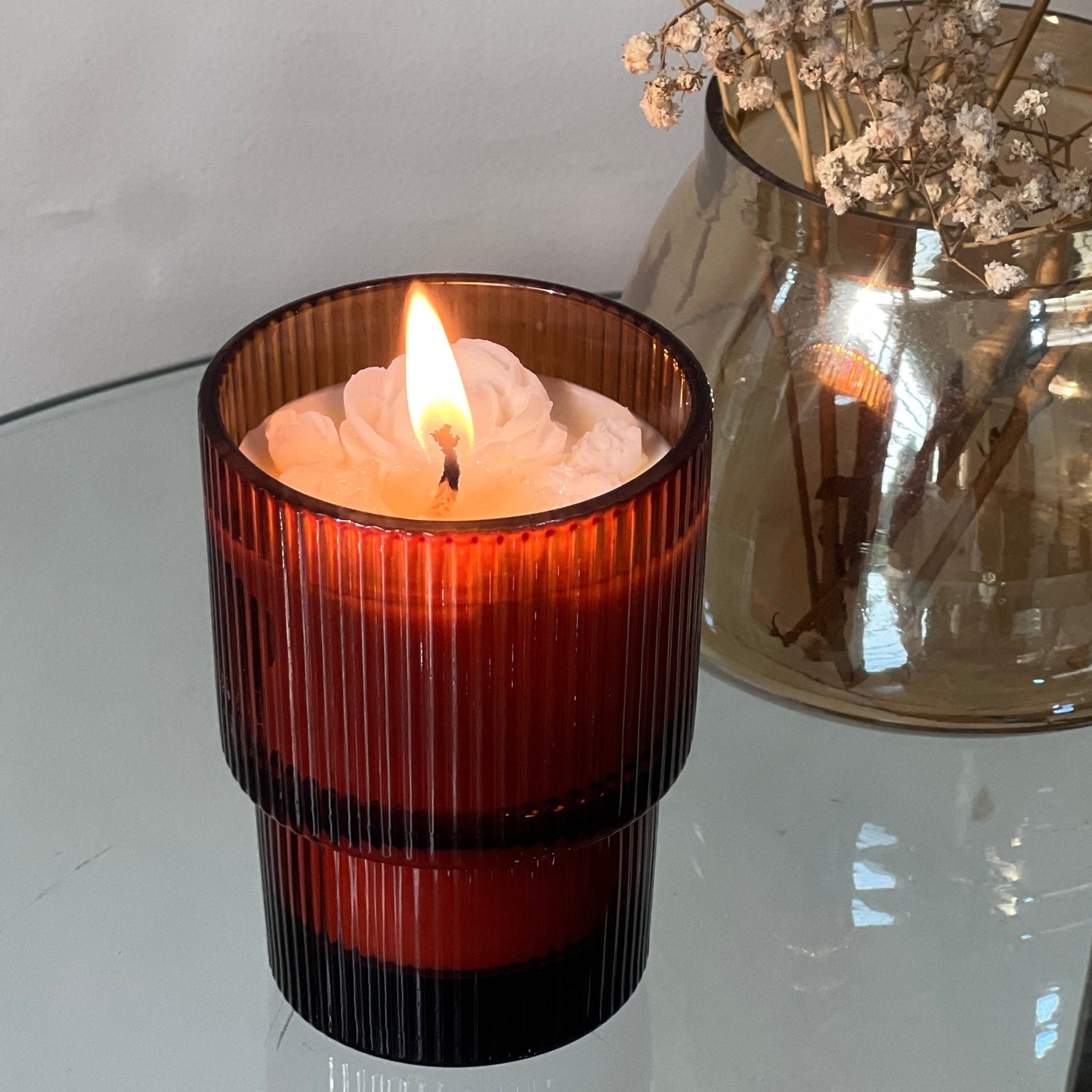 Indian vanilla candle in an amber ribbed glass jar with a vase of dried flowers on a reflective surface.