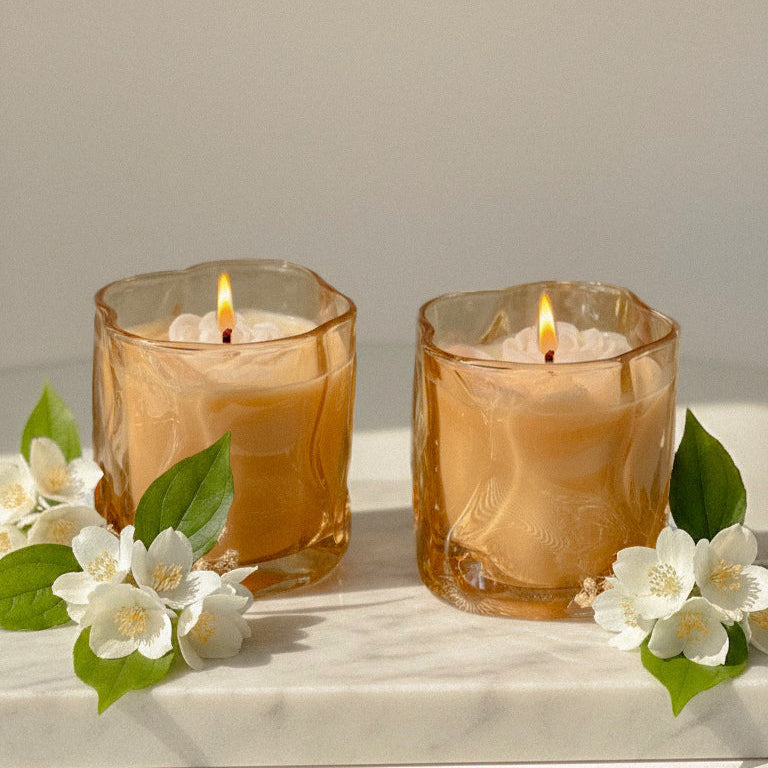 Two lit candles in golden glass holders with white flowers on a light surface.