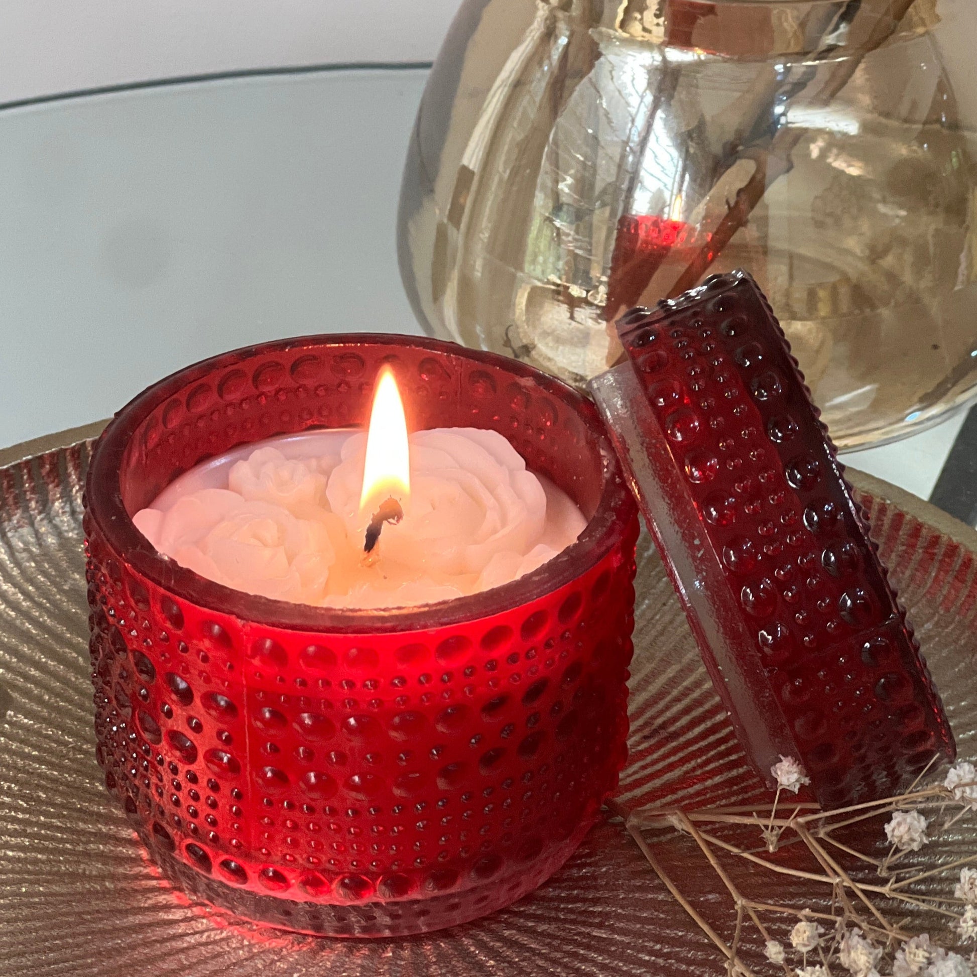 Candle in a red holder with a lit flame, surrounded by dried flowers and a metallic vase on a reflective surface.