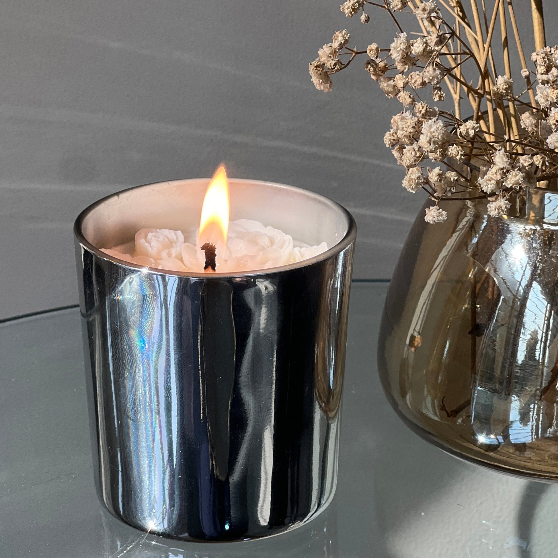 Silver candle holder with a lit candle and a vase of dried flowers on a reflective surface.
