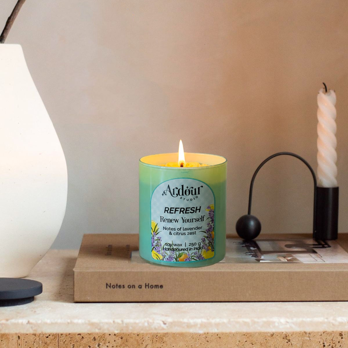 A lit candle in a decorative container on a surface with a lamp and magazine in the background.