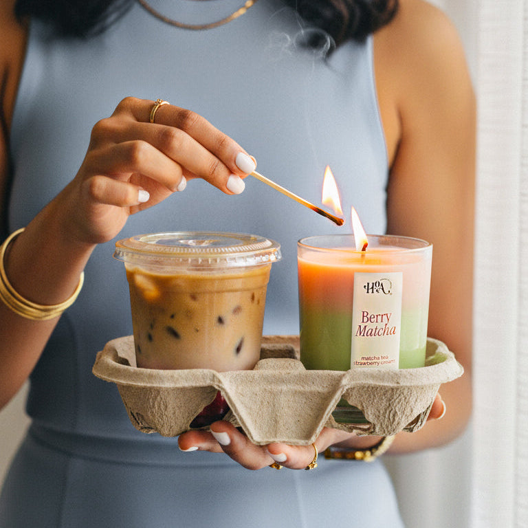 Woman lighting a berry matcha candle with a match, holding a cup of iced coffee in a carton.