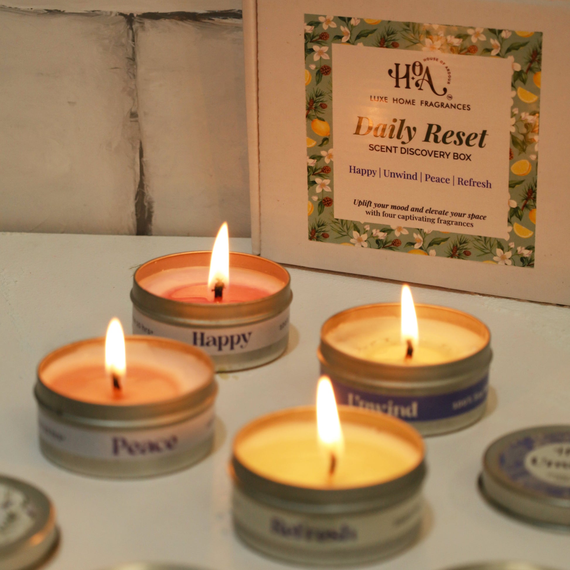 Candle tins with lit candles on a white surface, with a branded box in the background.