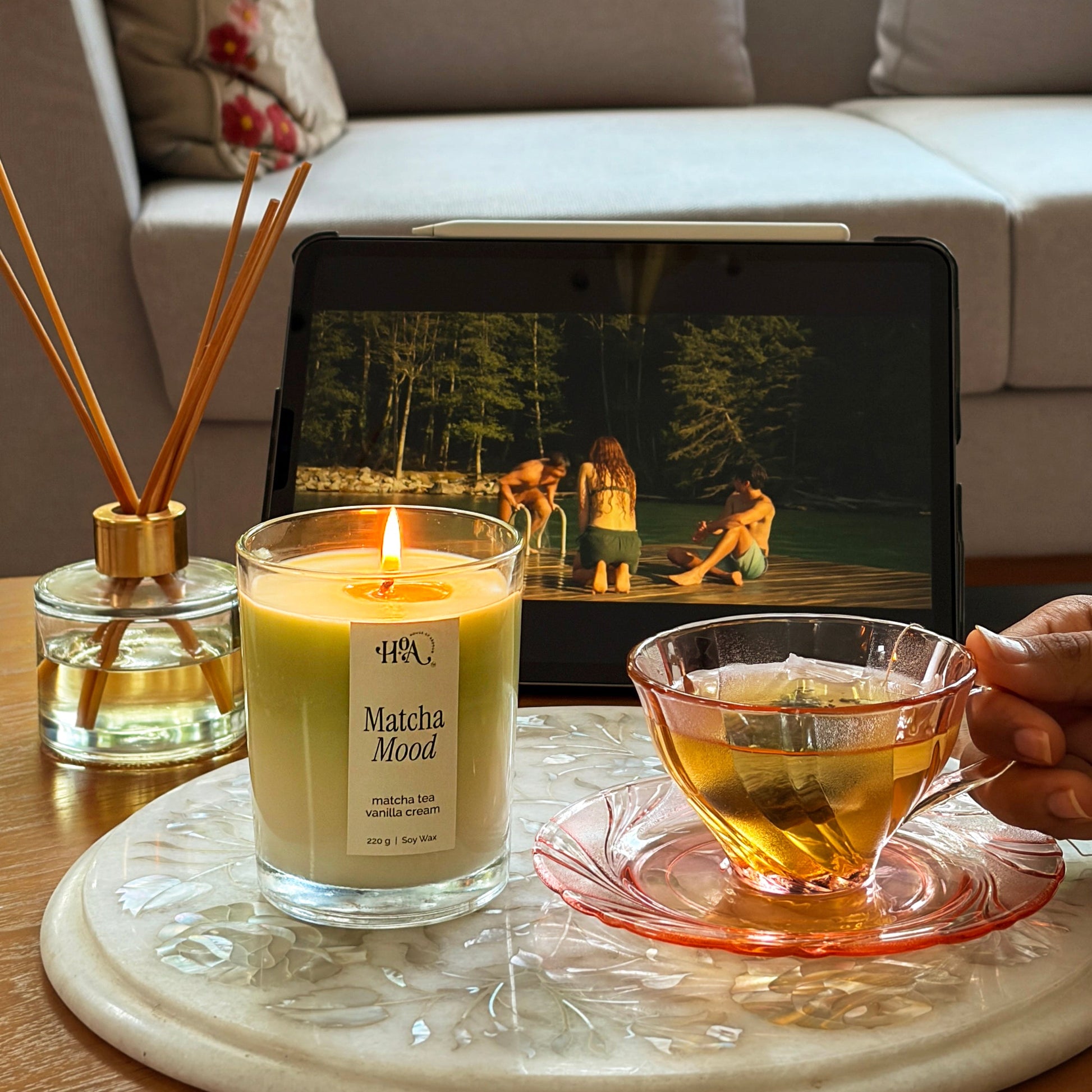 Candle, tea cup, and diffuser on a table with a laptop displaying a nature scene in a living room.