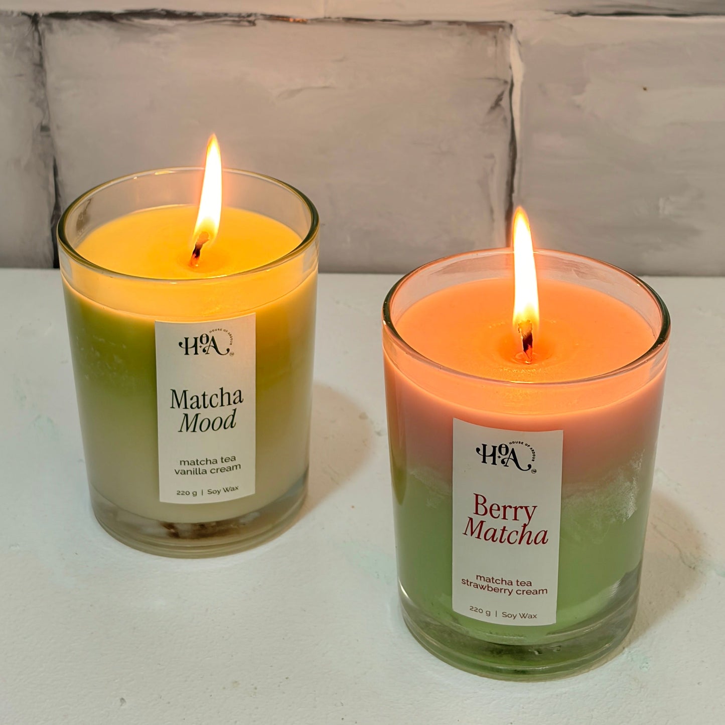 Two lit candles labeled 'Matcha Almond' and 'Berry Matcha' on a white surface with a light wooden background.