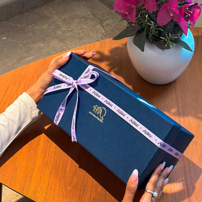 Blue gift box with a pink ribbon held by hands on a wooden table, next to a white vase with pink flowers.