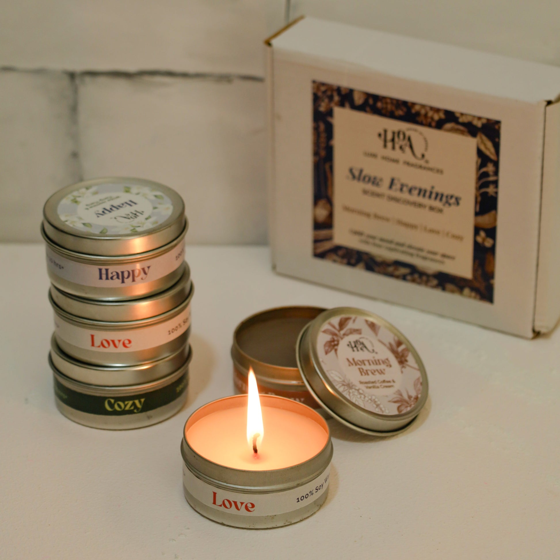 Stack of small candles with labels and a box labeled 'Slow Evening' on a white surface.