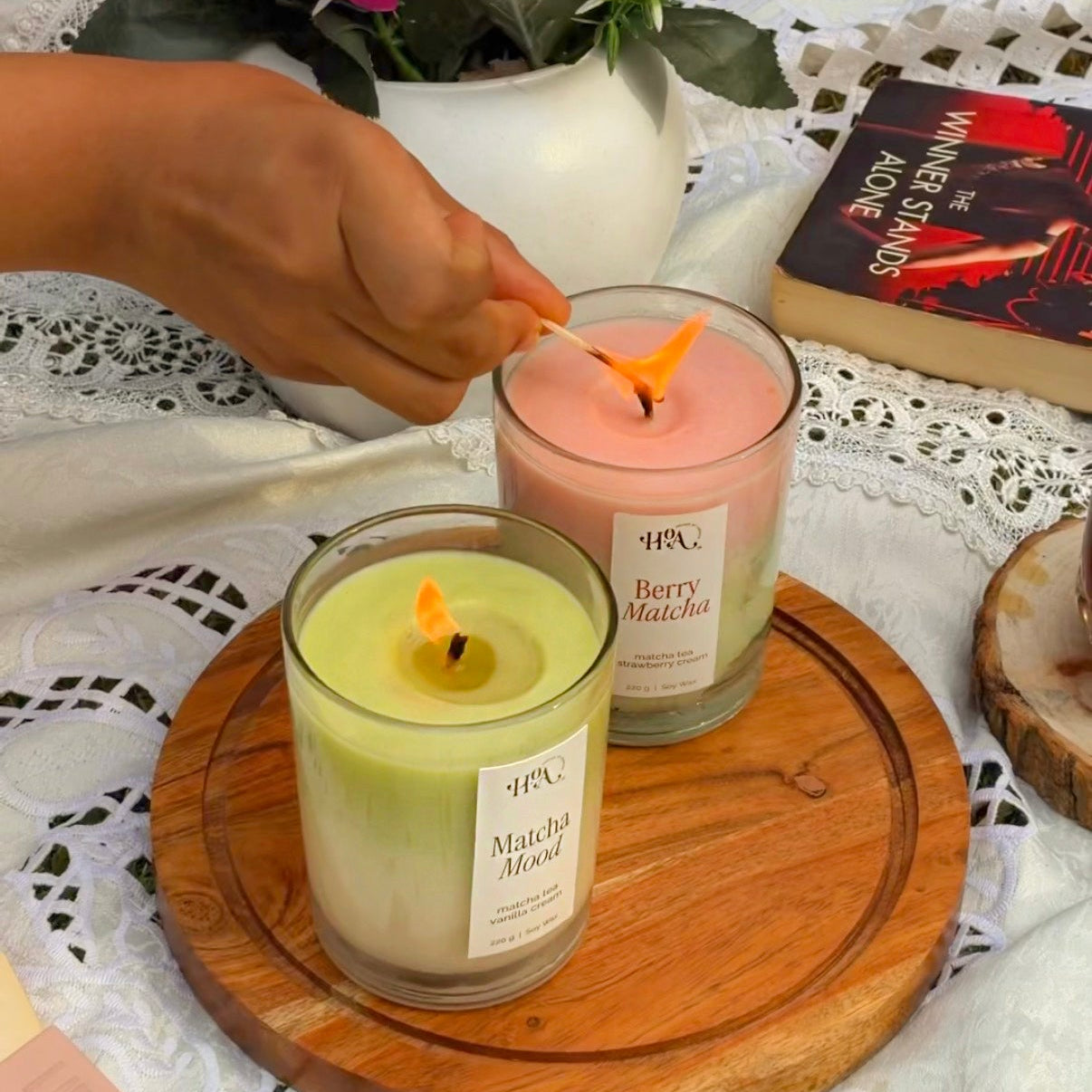 Two candles on a wooden coaster with a hand lighting one, a plant, and a book in the background.