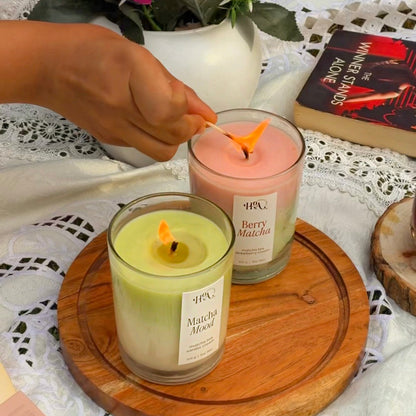 Two candles on a wooden coaster with a hand lighting one, a plant, and a book in the background.