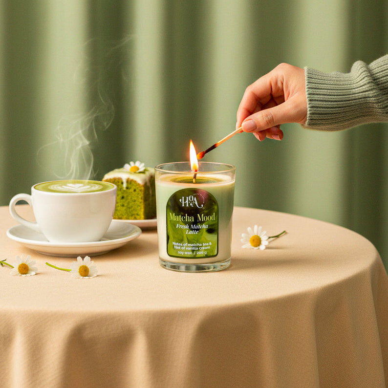Person lighting a candle with a cup of tea and a small cake on a table against a green curtain background.