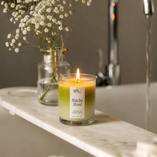 Candle labeled 'Matcha Mood' on a marble surface with a vase of flowers and a faucet in the background.