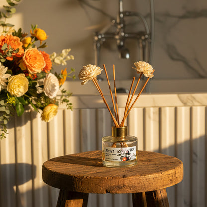 A small glass bottle with a floral diffuser on a wooden stool in a bathroom setting.