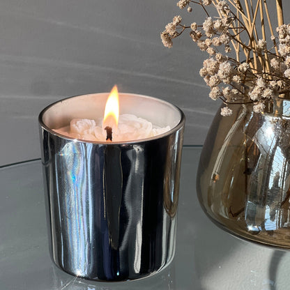 Silver candle holder with a lit candle and a vase of dried flowers on a reflective surface.